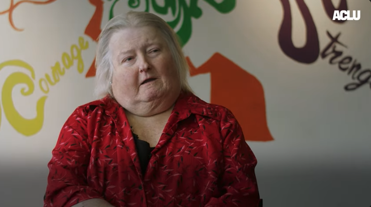 Aimee Stephens, a white transgender woman with silver hair wears a red dress and looks off to her left. She sits in front of a white wall.