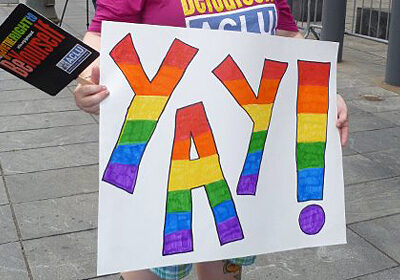 A person holding a sign reading "Yay!" in rainbow letters.