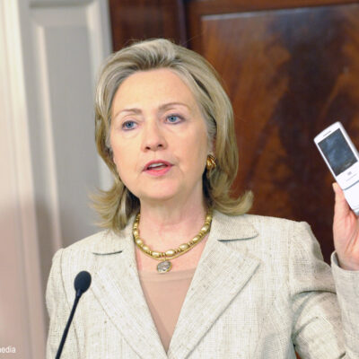 Hillary Clinton as Secretary of State at podium holding a cell phone