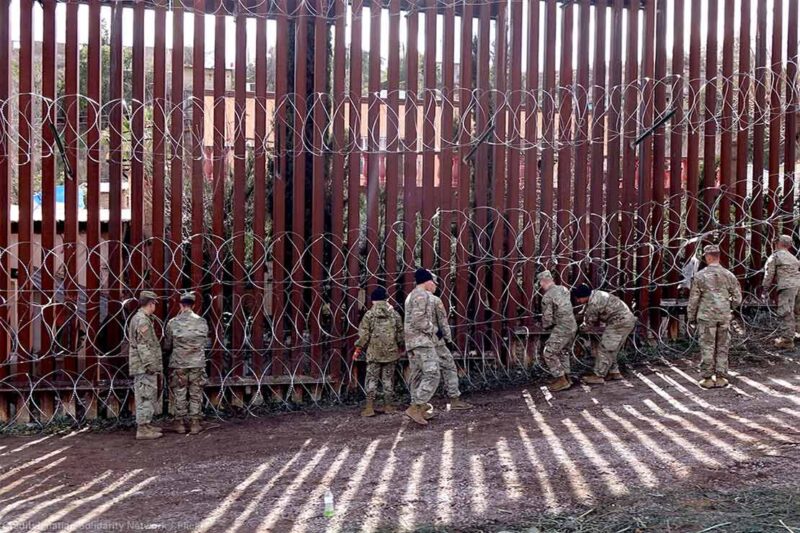 US military members are seen next to a red metal and barbed wire fence at the US border