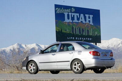 A silver car drives past the "Welcome to Utah" sign at the state border.