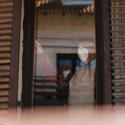An elderly person wearing a mask looks out a brown wood frame window.