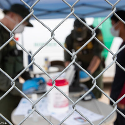 CBP agents wearing PPE processing someone wearing a mask behind a chain-link fence.