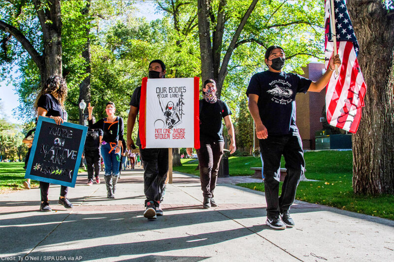 A group of people marching holding signs and a flag. The signs have text that read "You're on native land #landback" and "Our bodies our land / Not one more stolen sister".