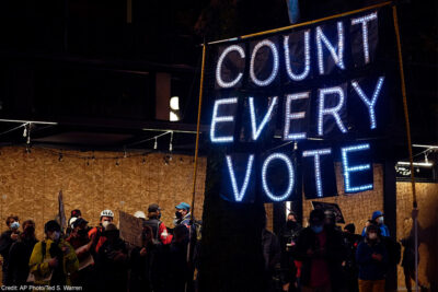 Demonstrators carrying a banner with the text "Count Every Vote" spelled out in LED bulbs.