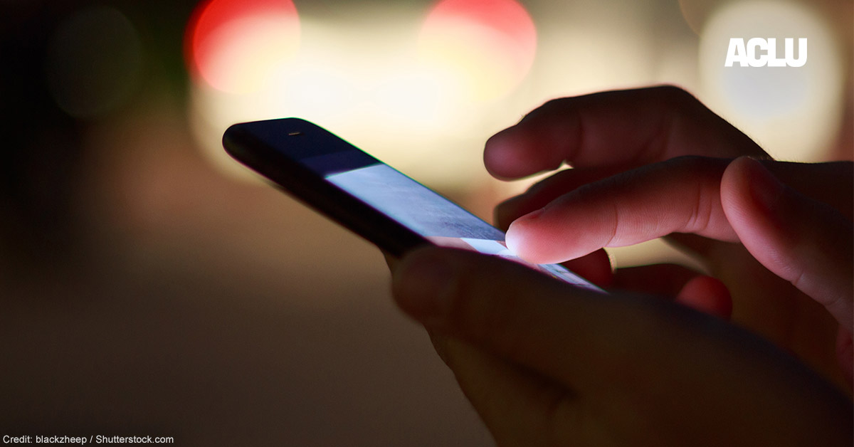 A pair of hands holding a cell phone at night with street lights in the background.