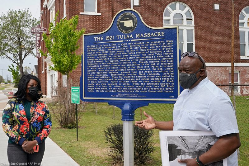 Two people explaining a sign that reads "the 1921 Tulsa Massacre" commemorating the original Black Wall Street