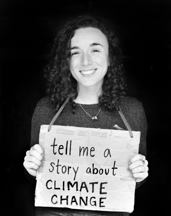 Black and white photo of Devi Lockwood holding a cardboard sign with the words "tell me a story about climate" on it.