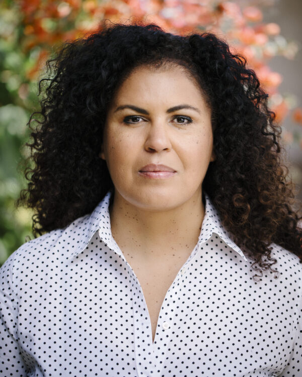 Headshot of Safiya Noble. Safiya Noble looks directly into the camera. She is wearing a white and black polka dot shirt and her background is blurred.