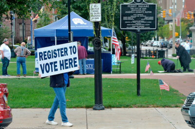 Person holds sign that reads "register to vote here" near a voter registration booth on a Pennsylvania street