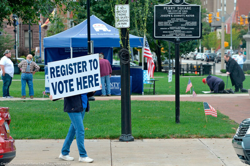 Person holds sign that reads "register to vote here" near a voter registration booth on a Pennsylvania street