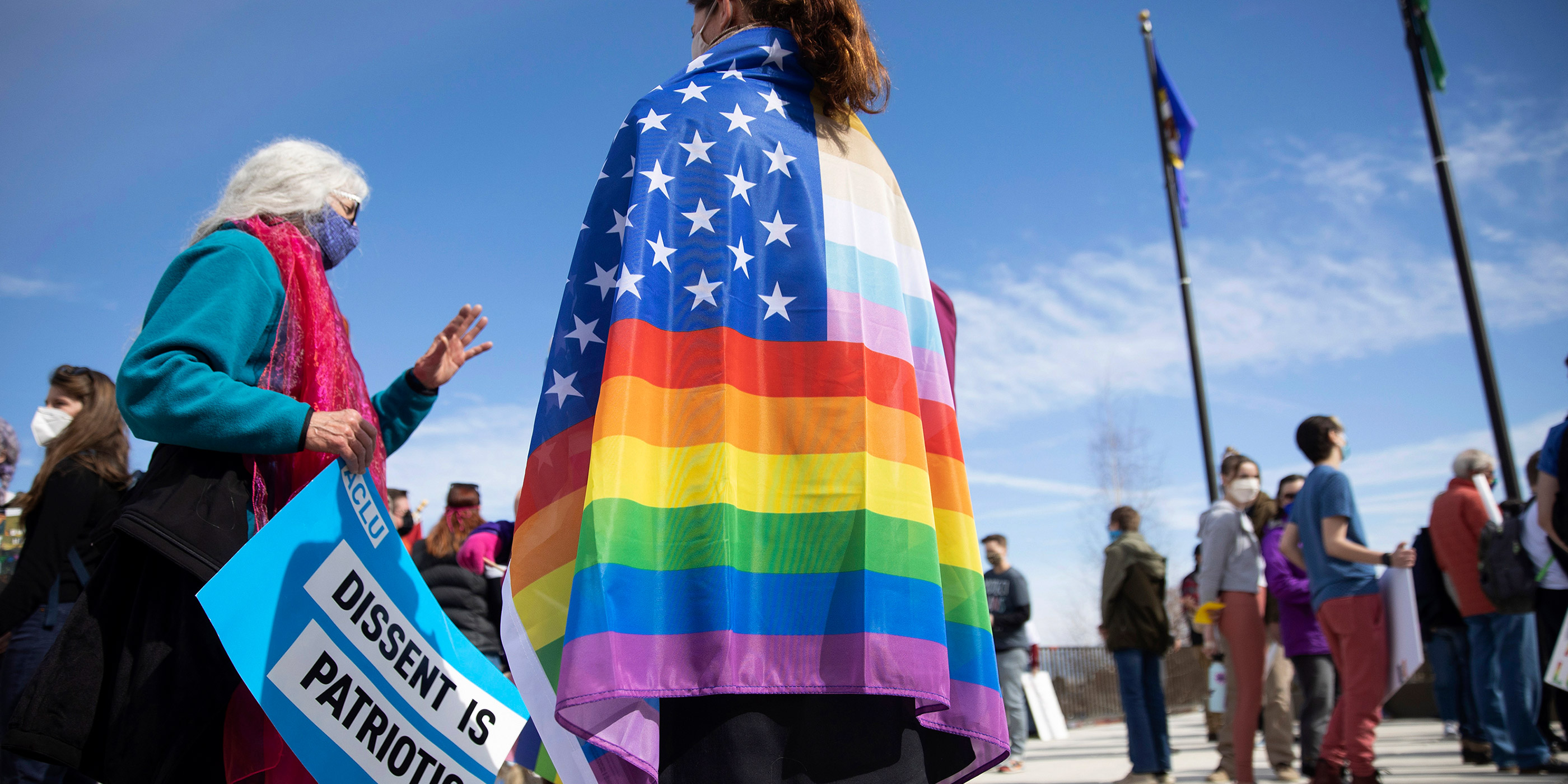 One demonstrator wears an LGBTQIA+ American flag while another holds a sign that says “Dissent is Patriotic.”