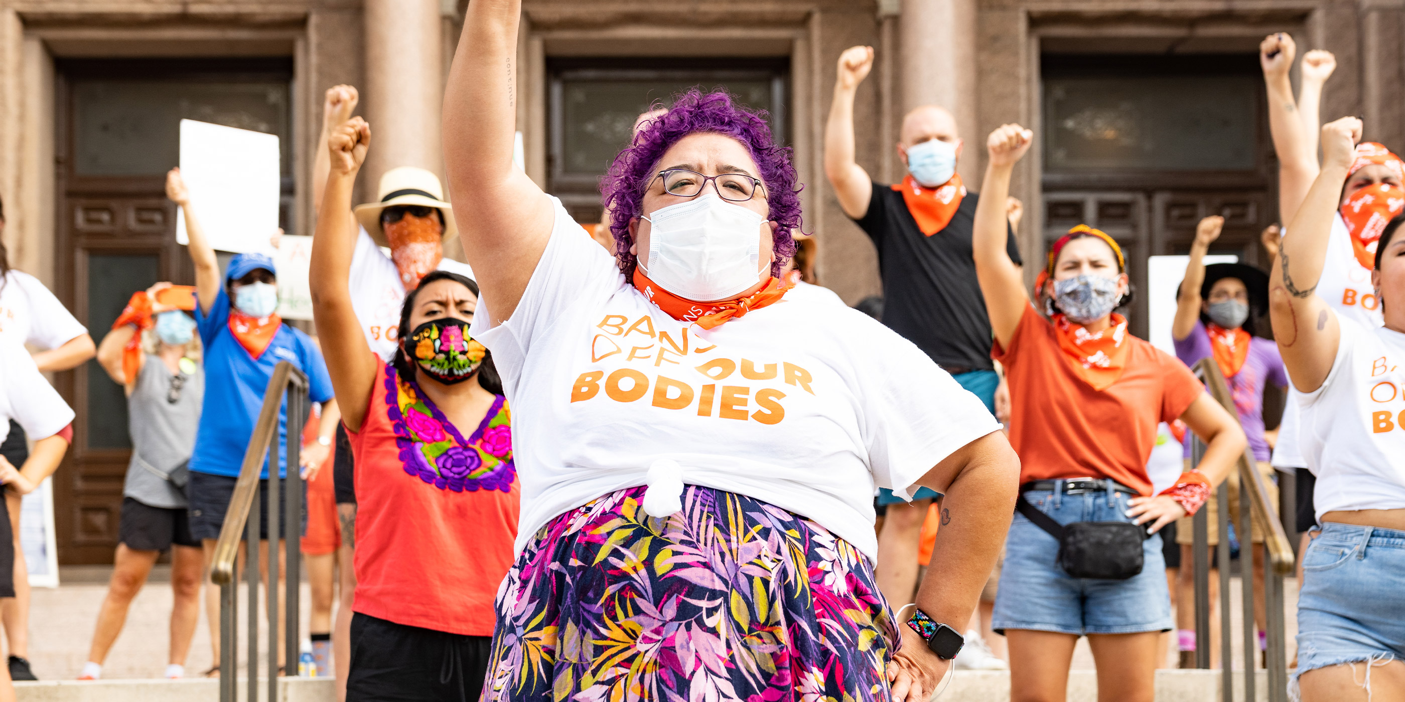 A group of demonstrators facing the camera with their fists in the air, with one wearing a shirt saying “Bans Off Our Bodies.”