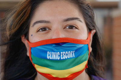 A close-up of Mariceli Alegria who's wearing a rainbow facemask with the printed words "Clinic Escort".