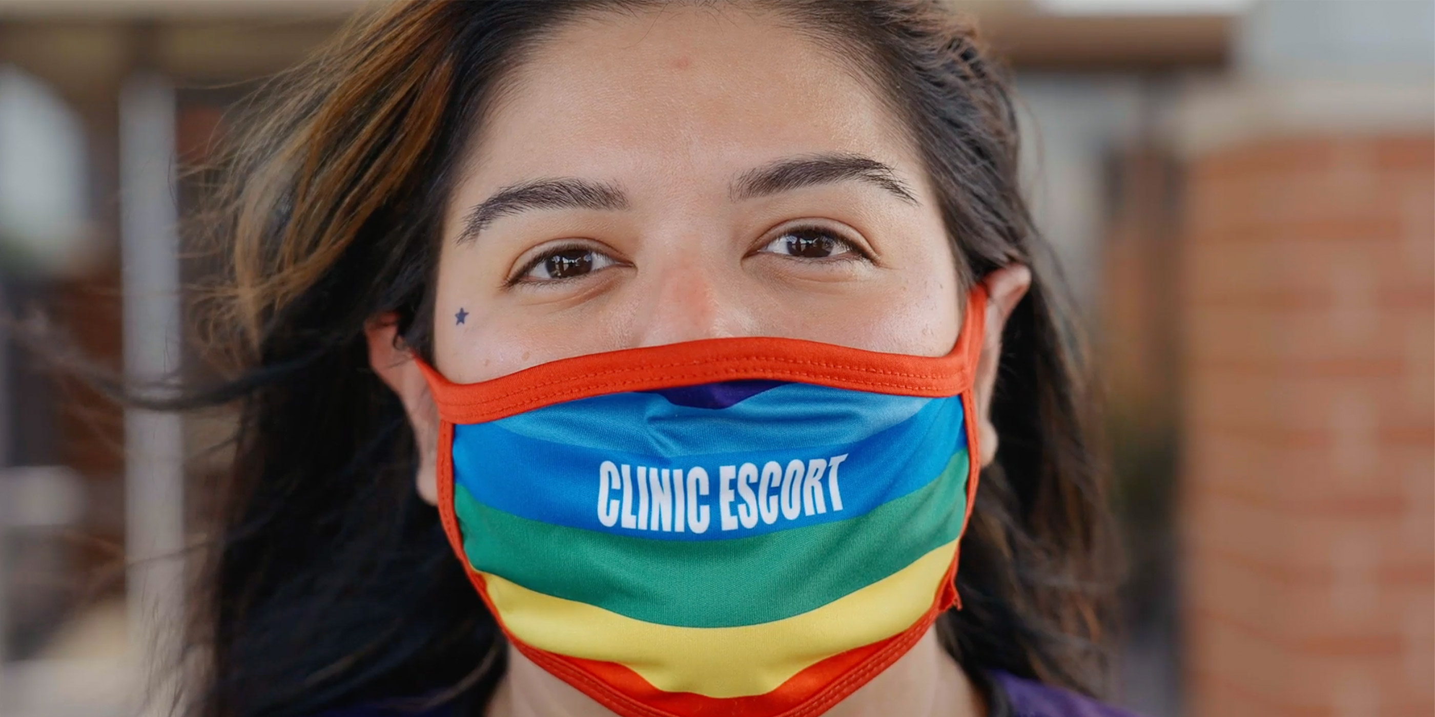 A close-up of Mariceli Alegria who's wearing a rainbow facemask with the printed words "Clinic Escort".