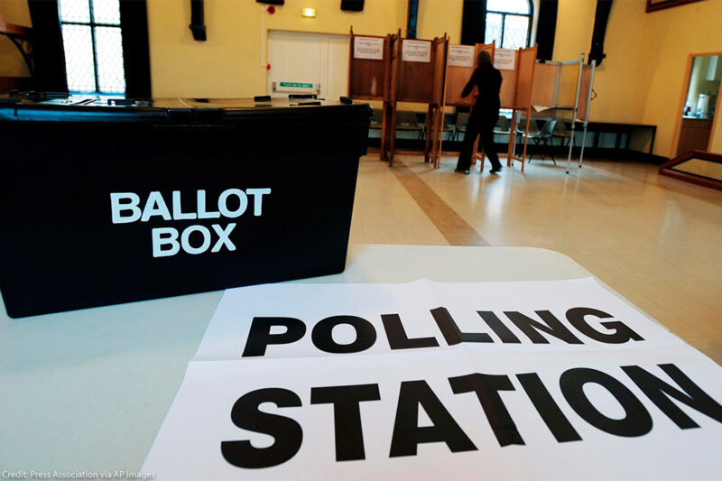 In the foreground, a black box with the words "BALLOT BOX" rests on a sign reading "POLLING STATION" at a polling station, while in the background a person in silhouette is at a voting booth.