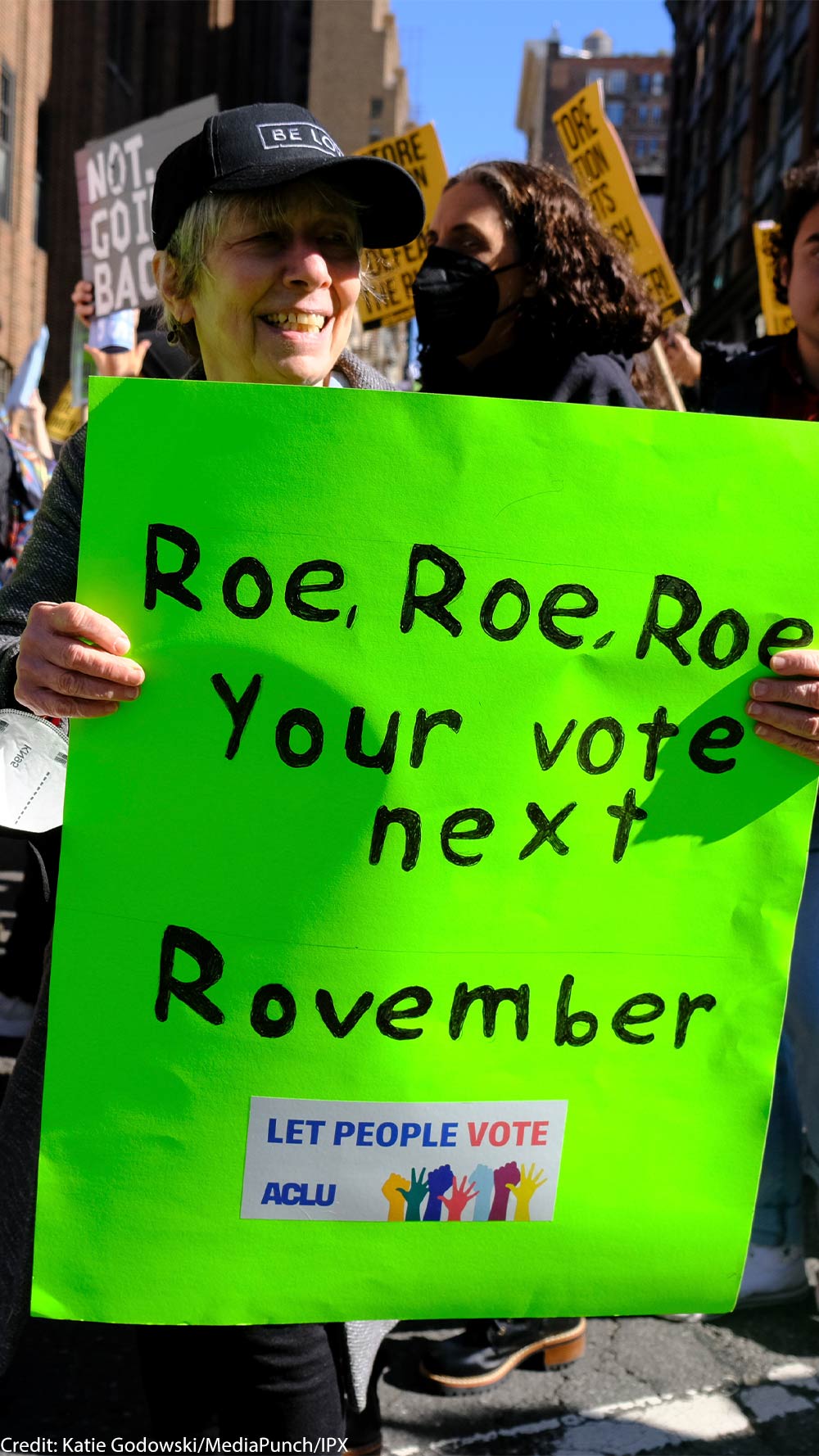 A woman holding a sign reading "Roe, Roe, Roe, Your vote next Rovember / LET PEOPLE VOTE ϰſ" poses for the camera as fellow activists walk behind her.