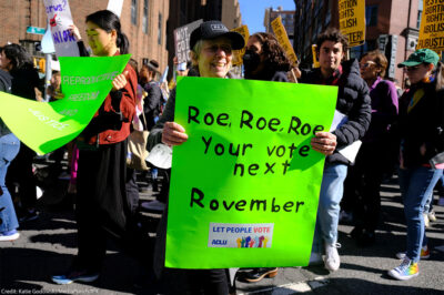 A woman holding a sign reading "Roe, Roe, Roe, Your vote next Rovember / LET PEOPLE VOTE ϰſ" poses for the camera as fellow activists walk behind her.