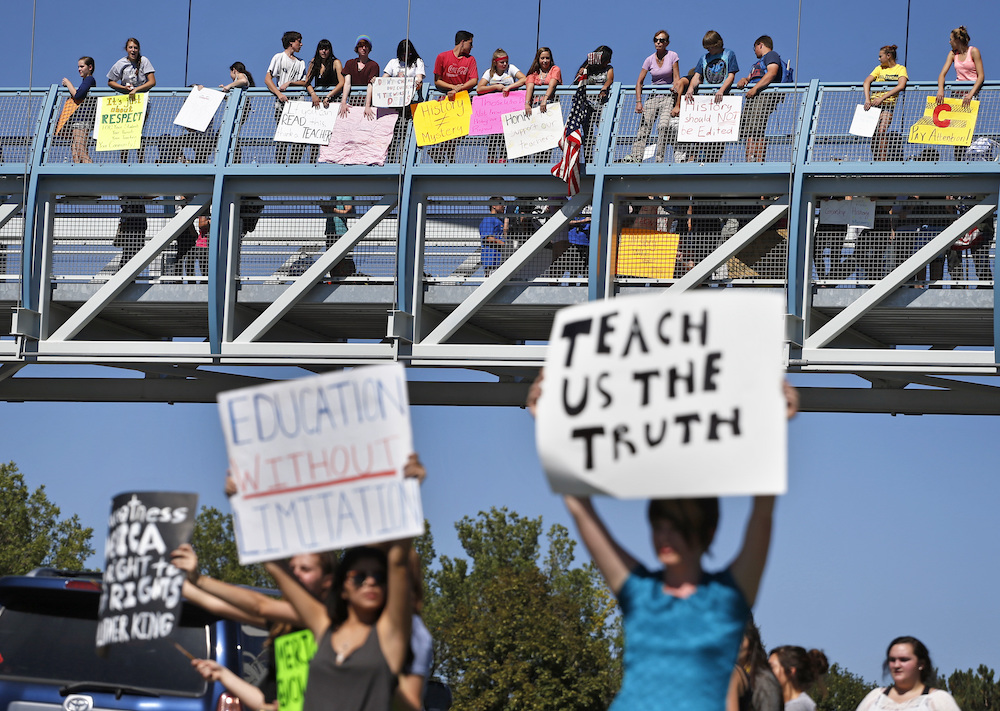 In the foreground (at an intersection,) two protesters carry signs with one reading "EDUCATION WITHOUT LIMITATION" and the other "TEACH US THE TRUTH", while in the background, other student demonstrators line an overpass protesting a proposal to emphasize patriotism and downplay civil unrest in the teaching of U.S. history.