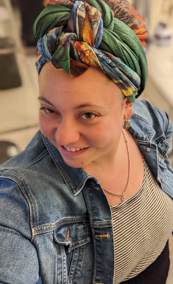 Headshot of Katherine smiling. She is wearing a multicolored scarf on her head, a denim jacket, and a black and white striped shirt.