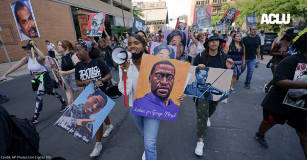 Protestors walking down the street, with one holding a microphone and a sign with art of George Floyd's face.