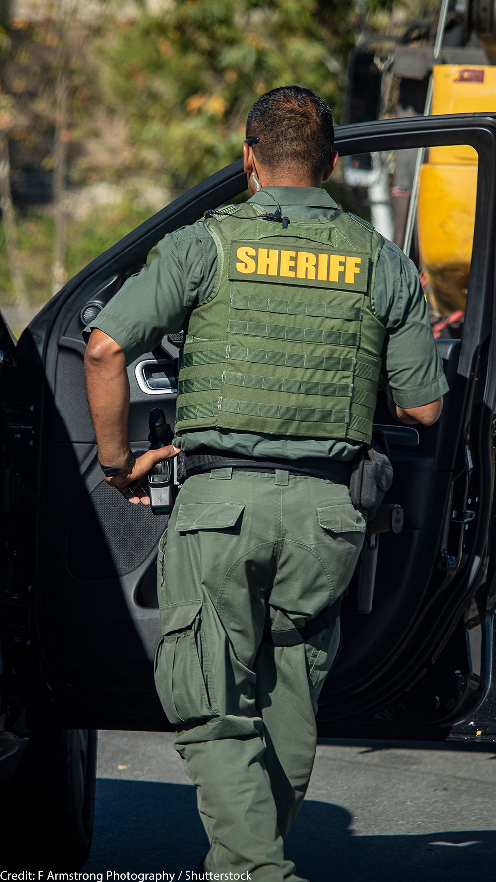 Shot of sheriff (wearing a bulletproof vest) from behind walking towards the open door of police car.