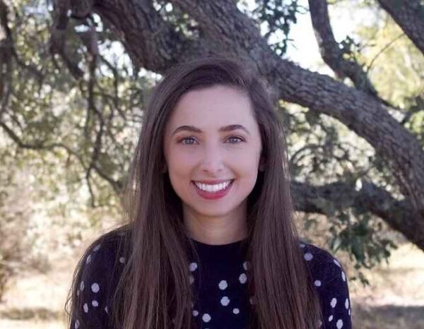 Headshot of ACLU of Texas attorney Chloe Kempf smiling. She's standing in front of a tree and wearing a Black sweater with white dots.