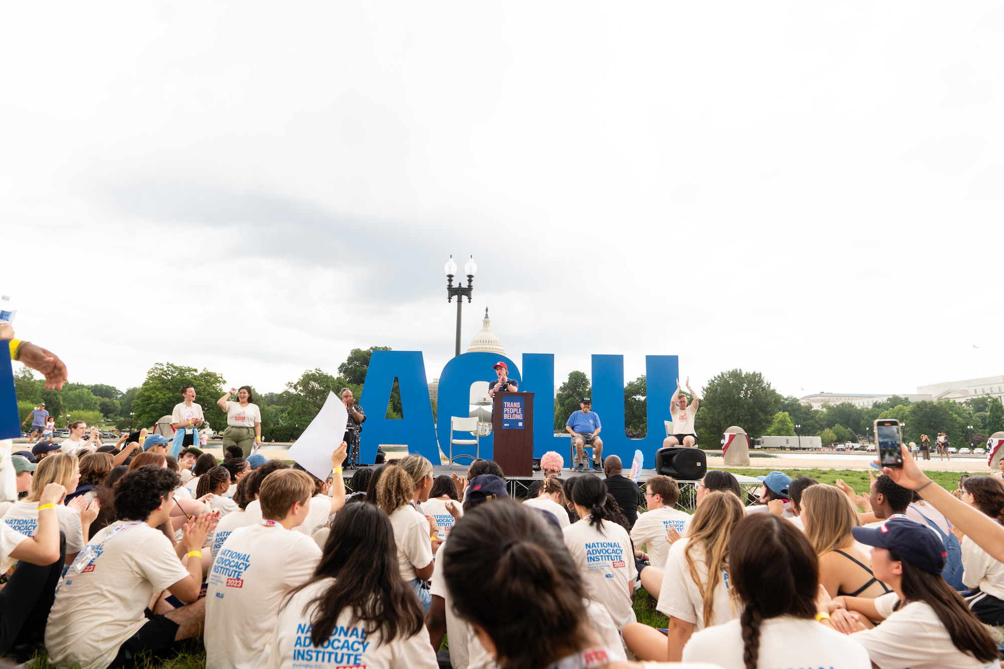 A speaker addressing a crowd in front of an ACLU sign and the U.S. capitol building.