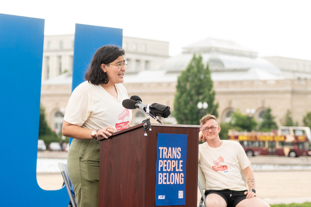 A speaker addressing a crowd in front of an ACLU sign and the U.S. capitol building.