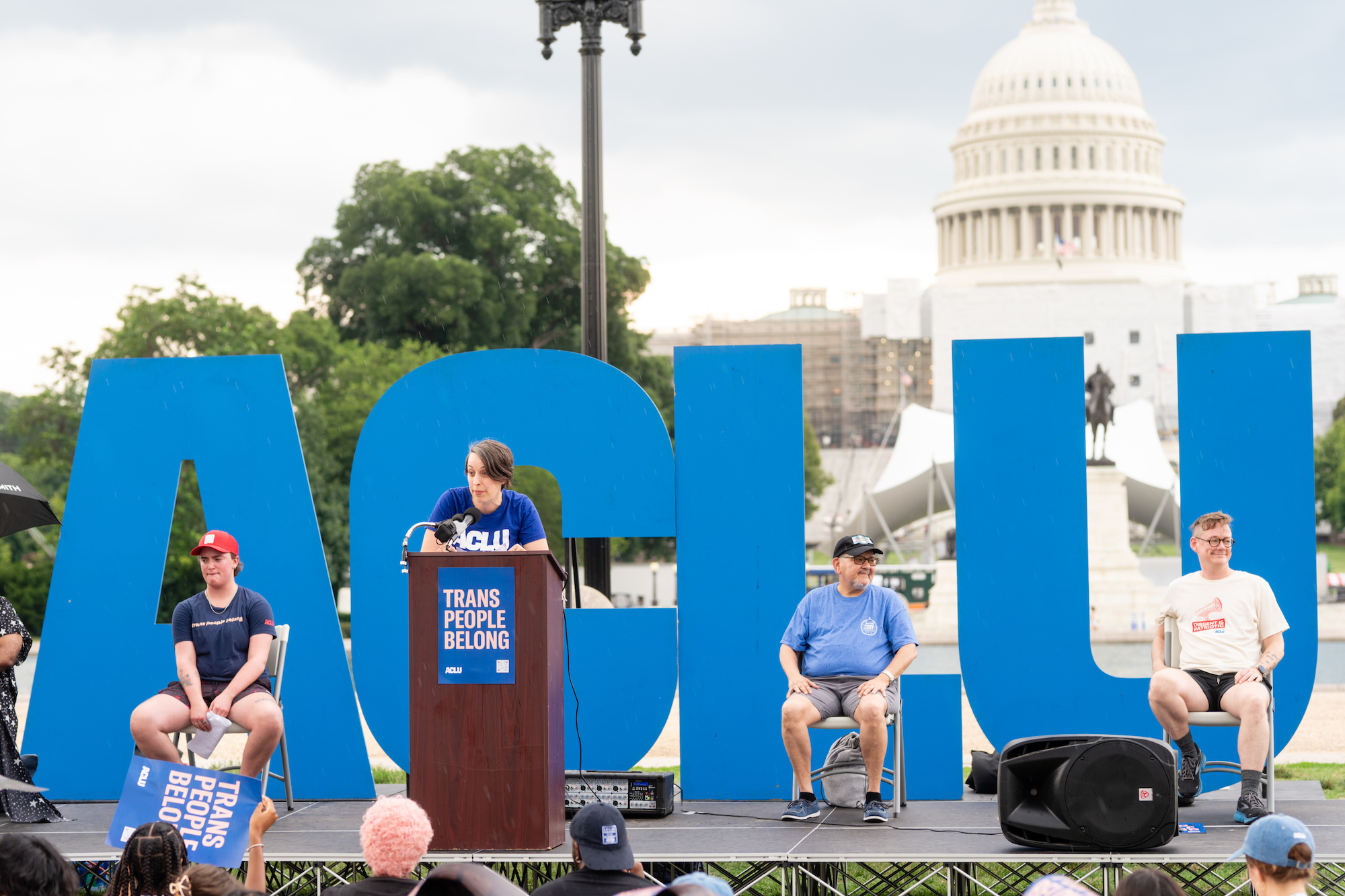 A speaker addressing a crowd in front of an ACLU sign and the U.S. capitol building.