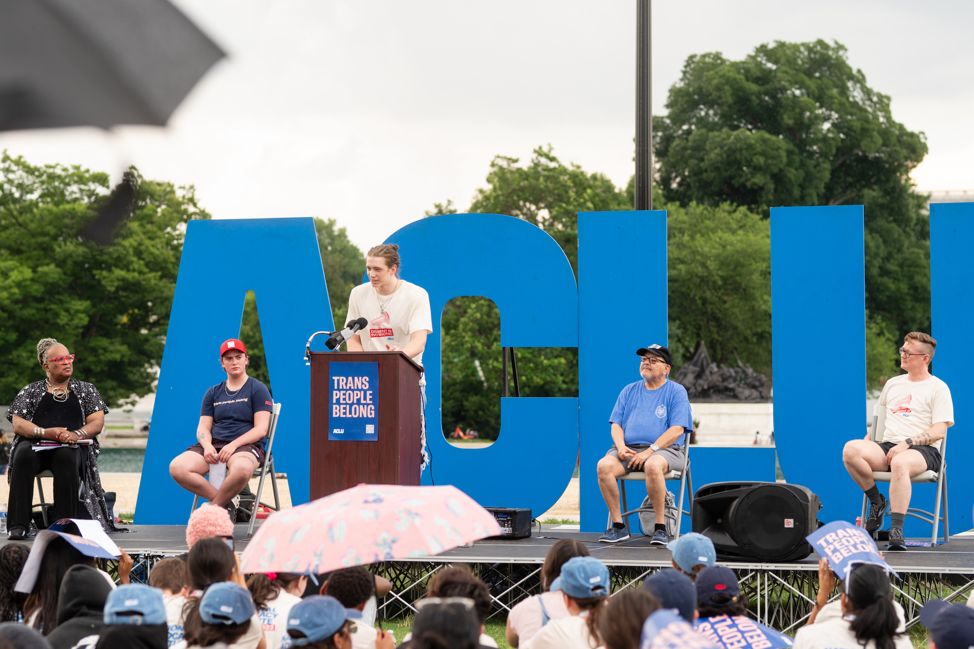 A speaker addressing a crowd in front of an ACLU sign and the U.S. capitol building.