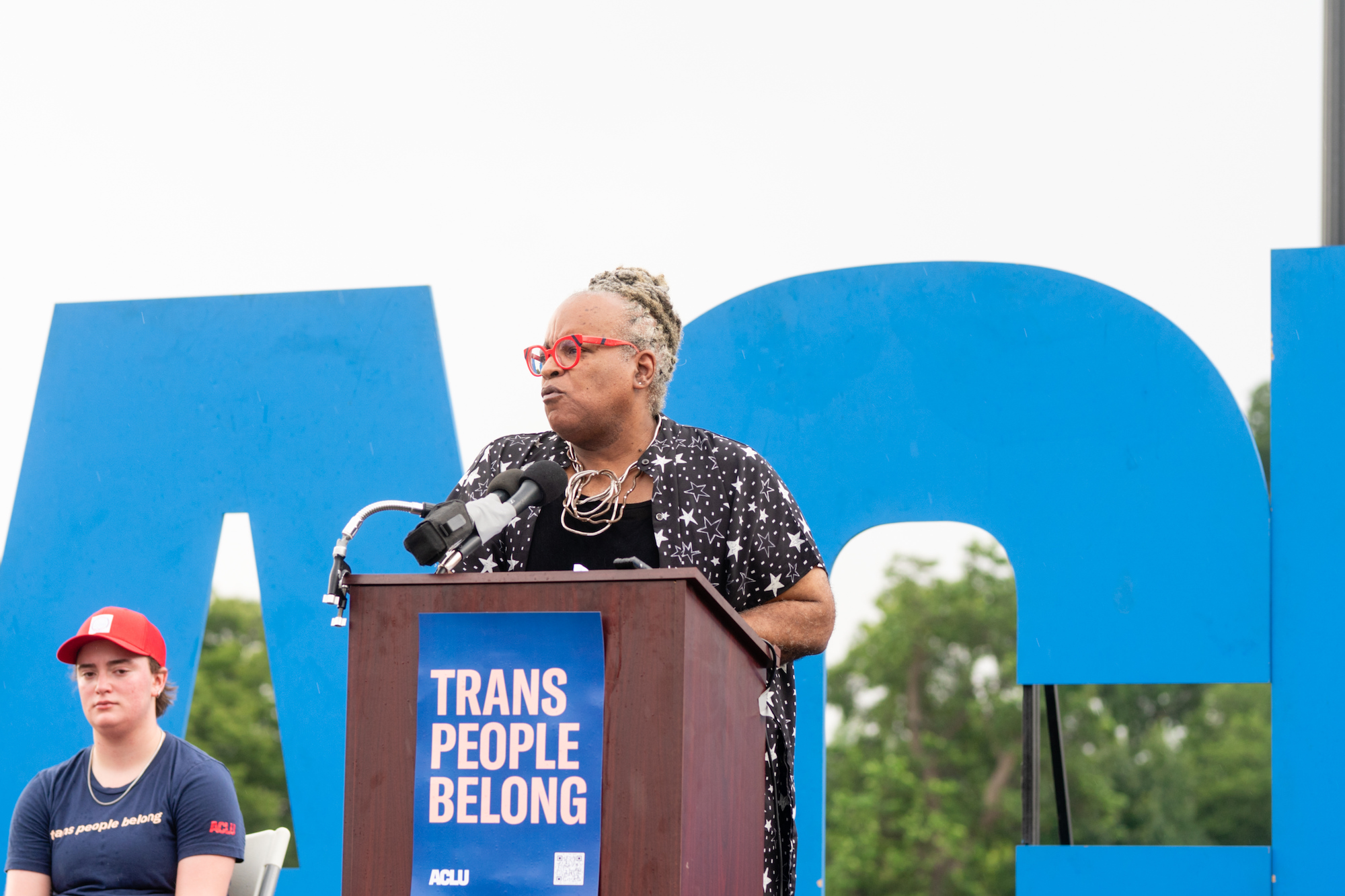 A speaker addressing a crowd in front of an ACLU sign and the U.S. capitol building.