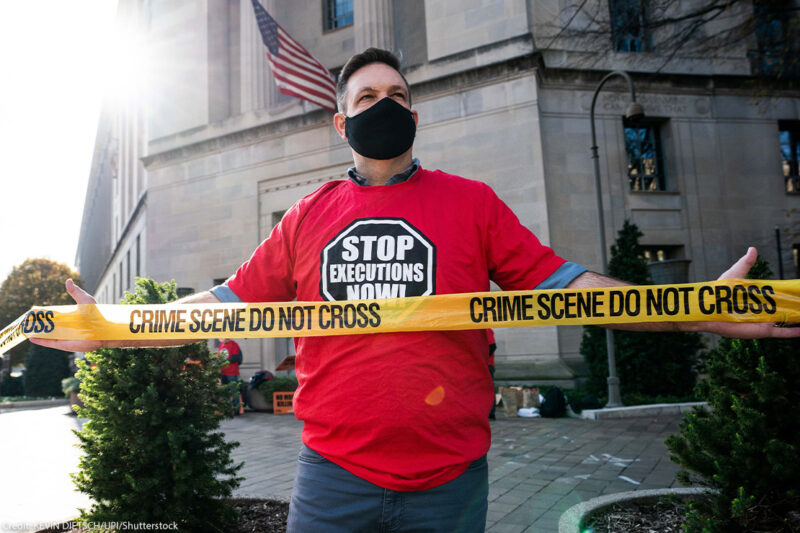 Man protesting against the death penalty in fron of the U.S. Justice Dept wearing a red shirt with a black stop sign reading "STOP EXECUTIONS NOW" in white letters.