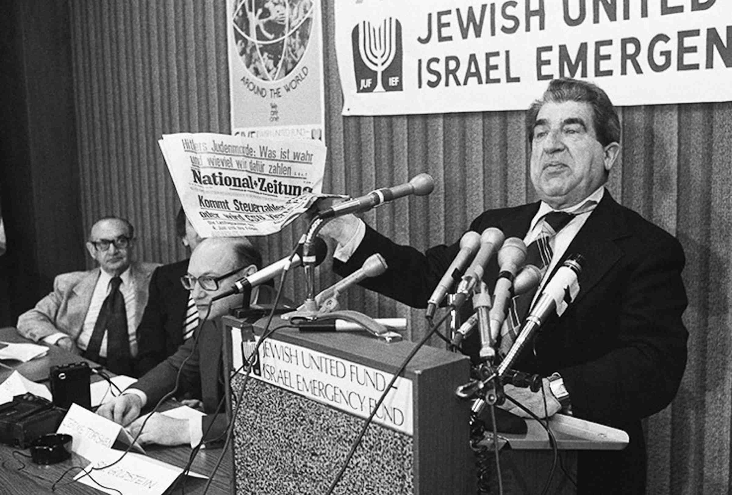 A black and white photo of a Sol Goldstein holding a newspaper at a podium with many microphones