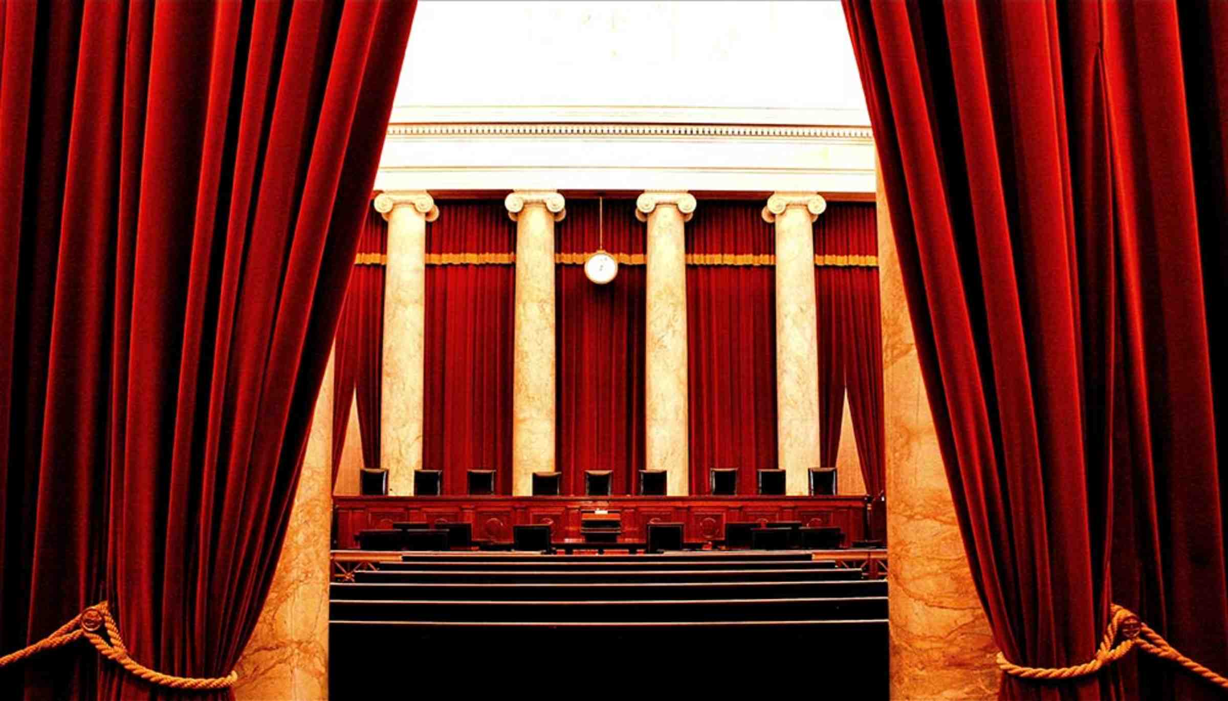 A look into the US Supreme Court through red curtains, featuring tall pillars and nine chairs.