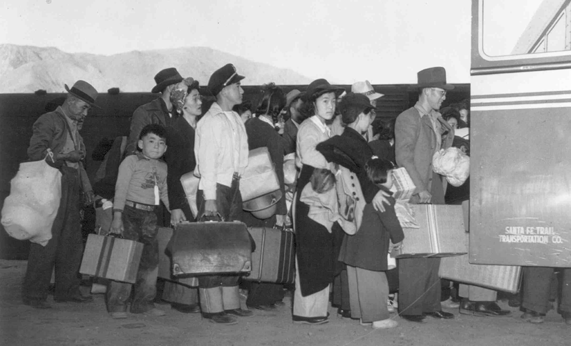 A black and white photo of a large group of Japanese-Americans holding luggage and waiting to board a bus