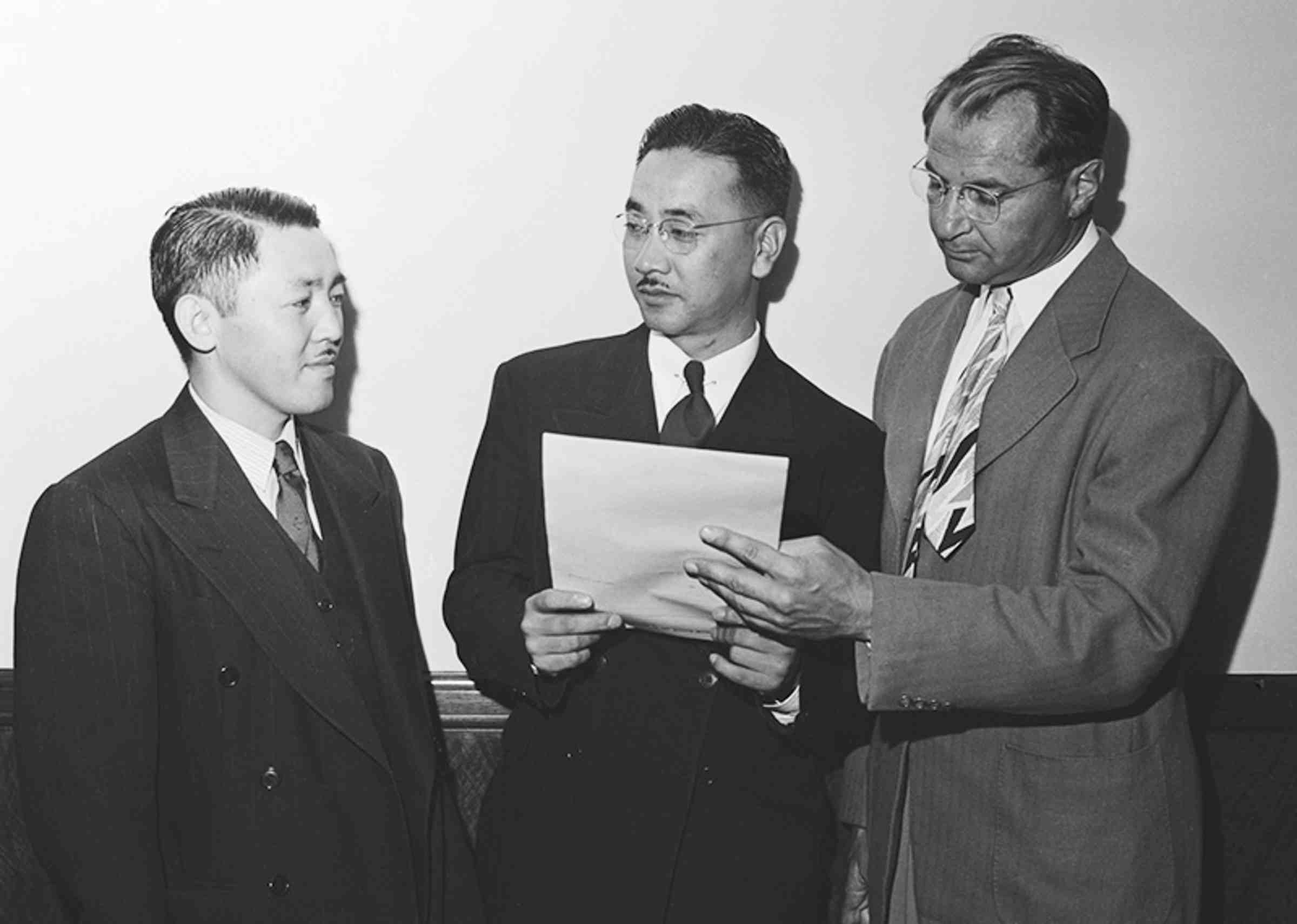 A black and white image of three people in suits looking at a piece of paper