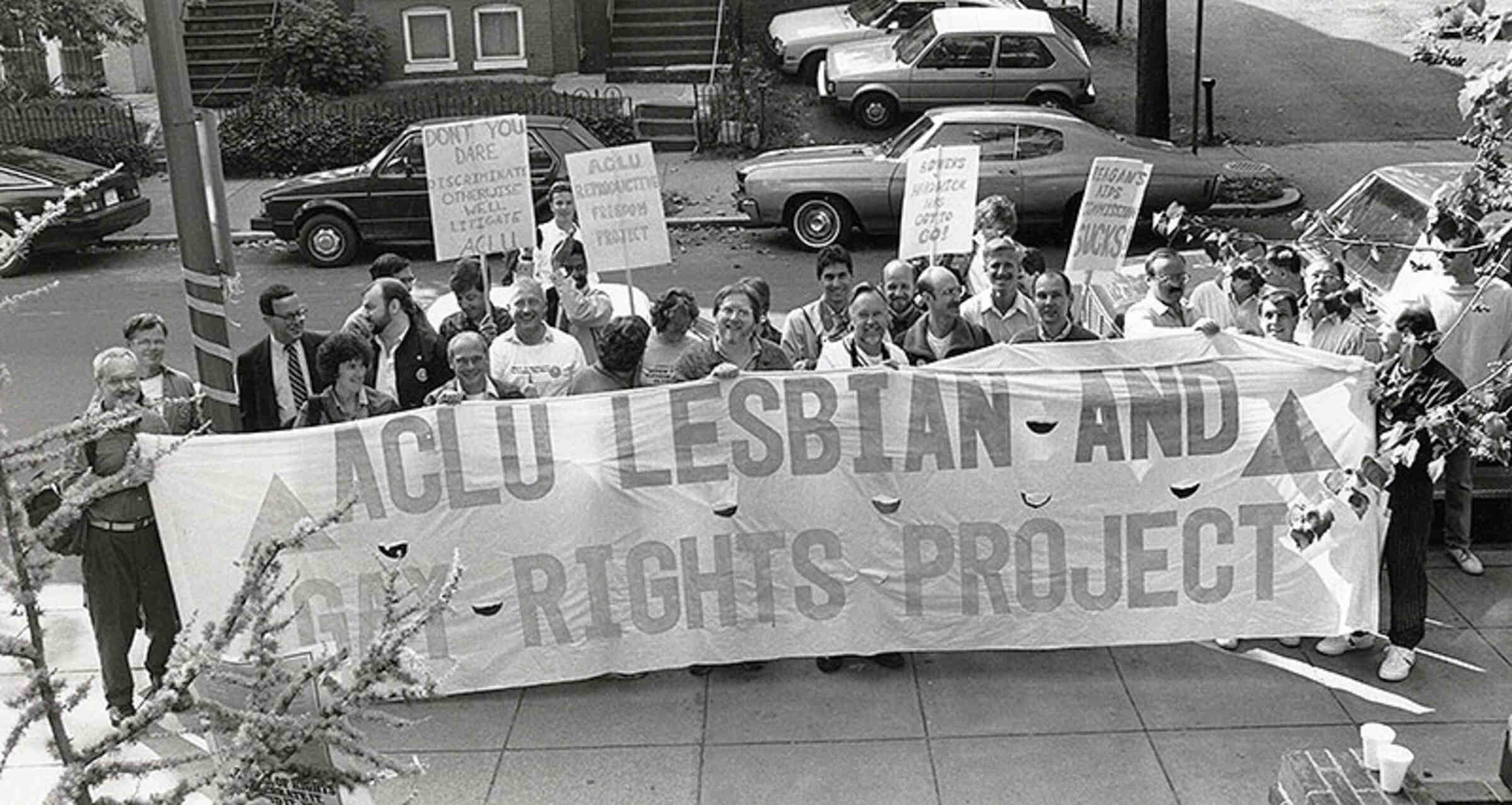 "ACLU LESBIAN AND GAY RIGHTS PROJECT" banner, seen before a march. Four individuals are holding signs that read "DON'T YOU DARE DISCRIMINATE OTHERWISE WE'LL LITIGATE ACLU," "ACLU REPRODUCTIVE FREEDOM PROJECT," "BOWERS V. HARDWICK HAS GOT TO GO!" and "REAGAN'S AIDS COMMISSION SUCKS!" 1987 (Credit: Tom Tyburski, 212-607-3300, 201-417-7298)