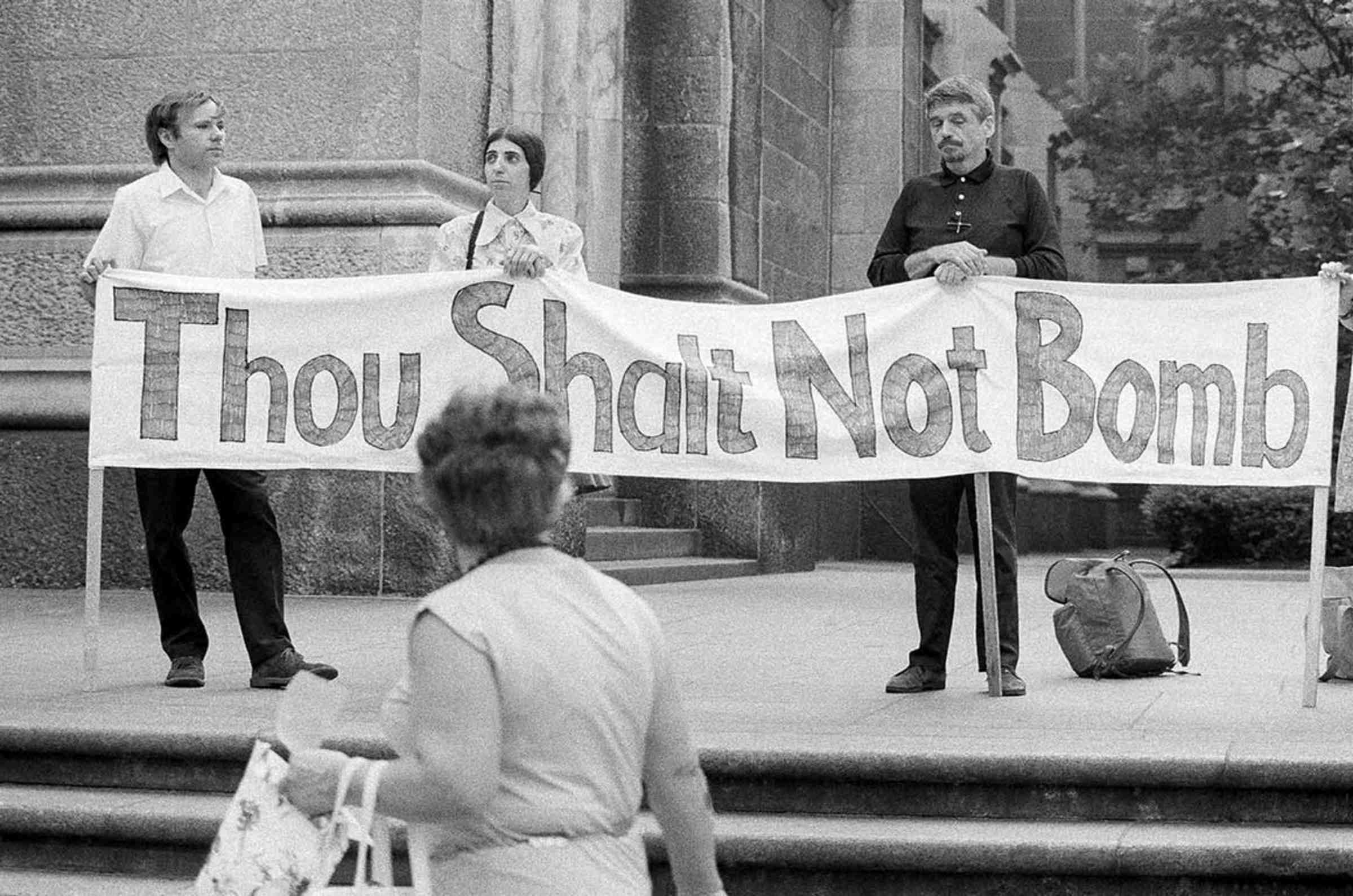 3 protestors holding a banner that says "Thou Shalt Not Bomb"