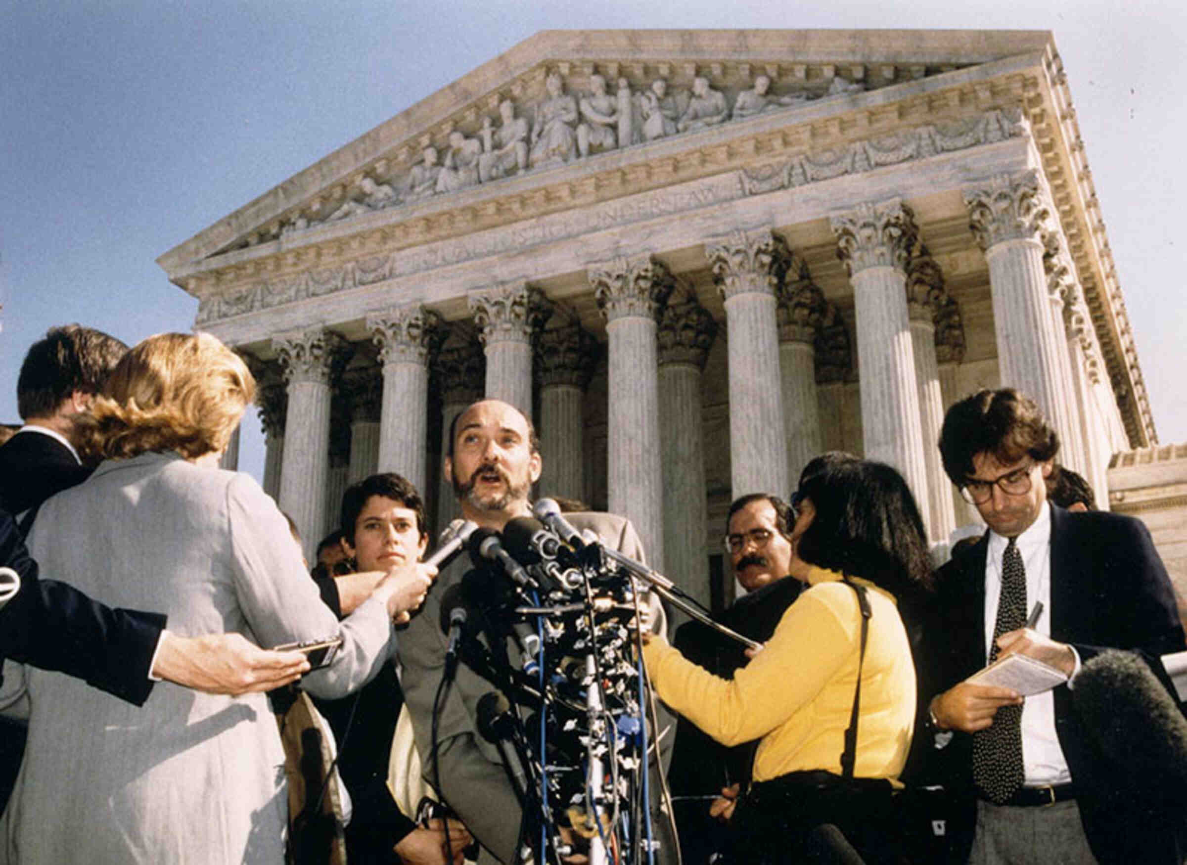 ACLU LGBT/AIDS Project Director Matt Coles speaking to the press after arguments in Romer v. Evans. Attorney Suzanne Goldberg stands behind him, 1996. (Credit: ACLU)