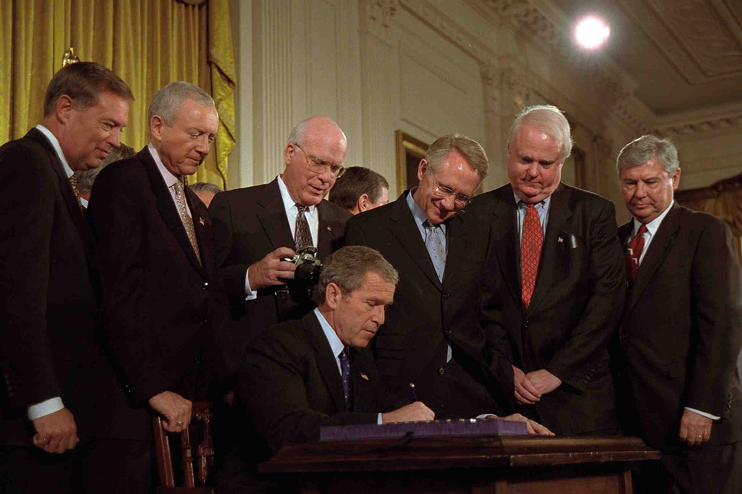 A group of men standing over George W. Bush as he signs a document