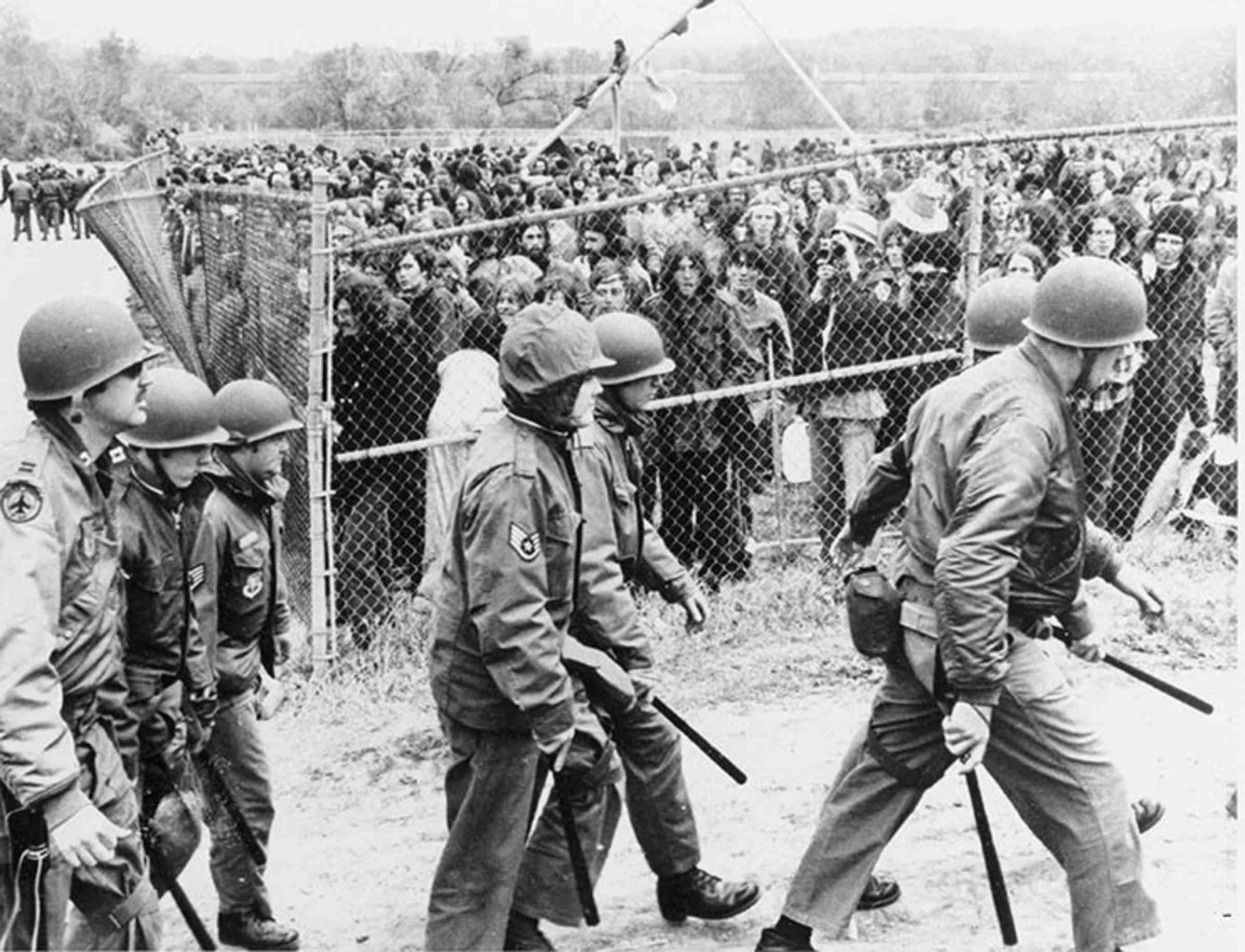 A large group of protesters stands behind a fence, with 7 officers walking by in the foreground