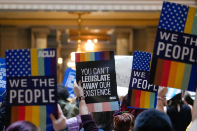 Individuals in a group holding ϰſ-branded signs saying "We the People," and "You Can't Legislate Away Our Existence."