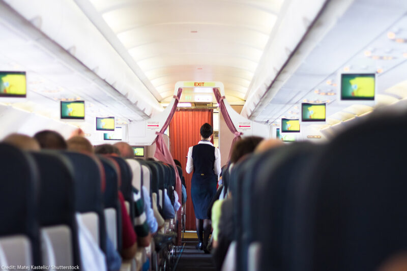 Interior of airplane with passengers on seats and flight attendant walking the aisle.