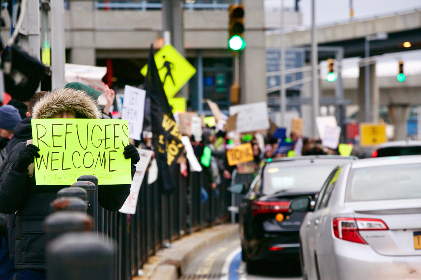 A crowd lined alongside a road, with the frontmost person holding a sign that says "REFUGEES WELCOME"