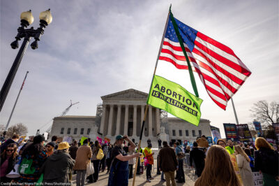 A demonstrator holds up a pole with an American flag on top and another flag reading "ABORTION IS HEALTHCARE" below it as others protest the proposed limited use of mifepristone outside the U.S. Supreme Court on the 26th of March 2024.