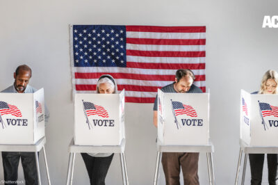 With and American flag in the background, four people in the act of voting, stand behind voter booths as they make their selections.