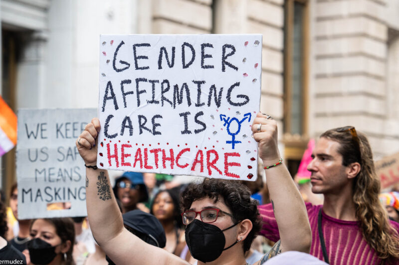 A shirtless demonstrator (wearing glasses and a black surgical mask) holds up a sign that reads" GENDER AFFIRMING CARE IS HEALTHCARE".