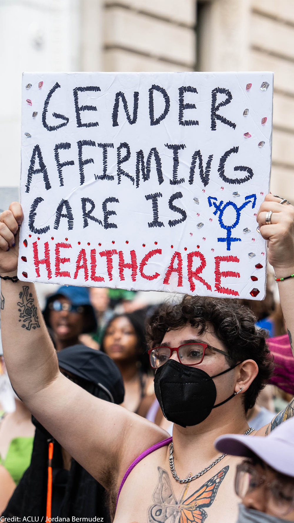 A shirtless demonstrator (wearing glasses and a black surgical mask) holds up a sign that reads" GENDER AFFIRMING CARE IS HEALTHCARE".