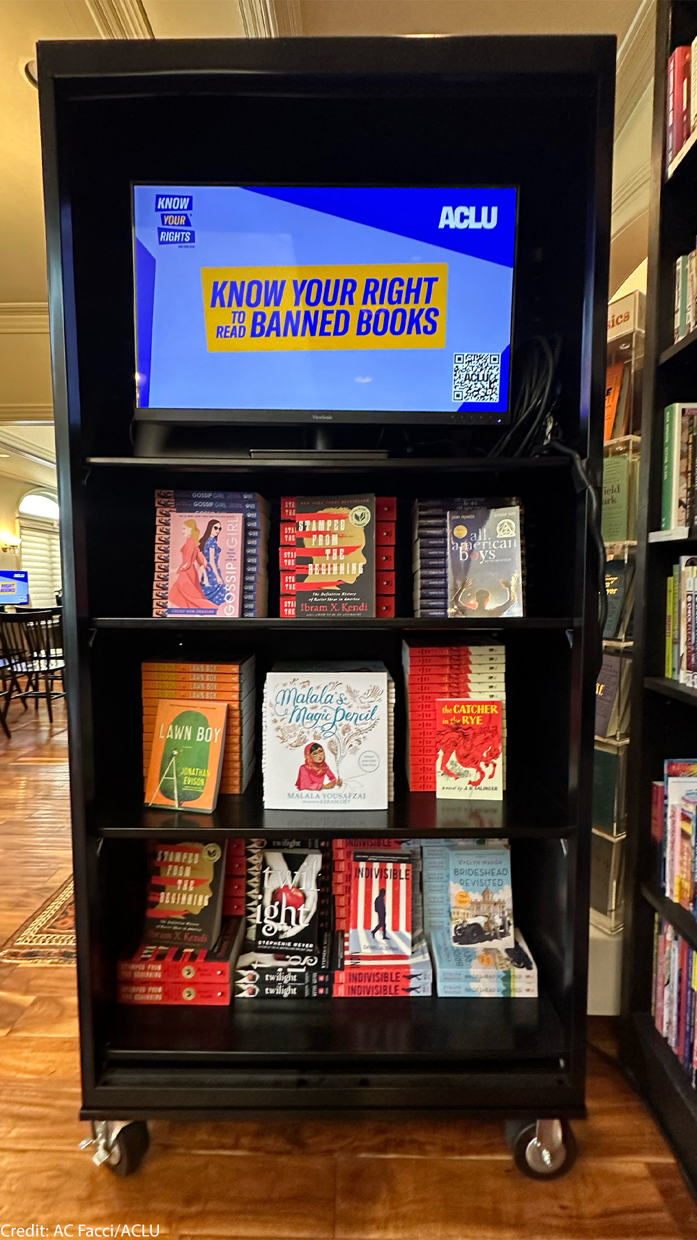 A black bookshelf on wheels that has a sign reading "KNOW YOUR RIGHT TO READ BANNED BOOKS"on the top shelf and a slew of banned books on the 3 lower shelves.